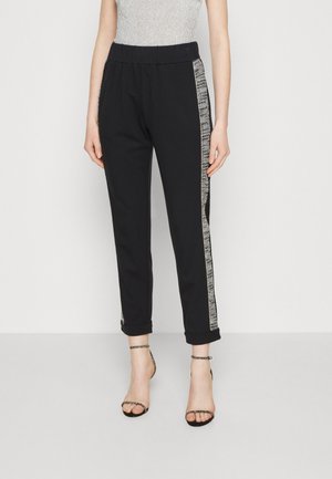 Mujer con pantalones negros ajustados en el tobillo con franjas laterales de lentejuelas plateadas y sandalias negras de tacón alto con pulsera en el tobillo, de pie frente a un fondo blanco.