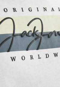 Bílý tričko se šedými a světle zelenými vodorovnými pruhy, s vyvýšeným černým potiskem "Jack Jones" a "ORIGINAL WORLDWIDE" vytisknutým pod ním.