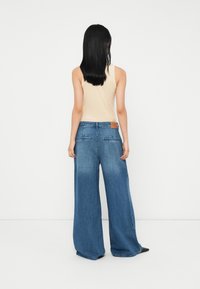 Weite blaue Denim-Jeans mit mittelhohem Bund, ausgestattet mit einem braunen Lederpatch auf der Rückseite und zwei Gesäßtaschen.