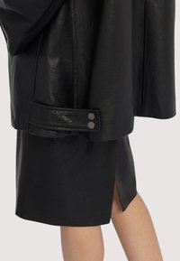 Svart läderjacka med en mjuk textur, utrustad med en vikbar krage, två metalliska tryckknappar och en sida-springa kjol i matchande tyg.