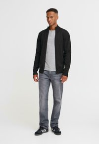 En ung man står självsäkert, iklädd en svart zip-up jacka, grå t-shirt och blåjeans, framför en vanlig vit bakgrund.