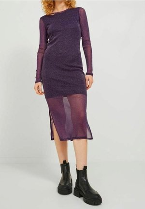 Mujer con un vestido midi morado translúcido de manga larga con una abertura, combinado con botas negras gruesas hasta el tobillo, sobre un fondo liso.