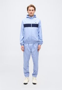 Νέος άνδρας που στέκεται φορώντας ένα ανοιχτό γαλάζιο φόρμα με ένα ναυτικό ρίγα στο στήθος και ανοιχτόχρωμα αθλητικά παπούτσια σε λευκό φόντο.