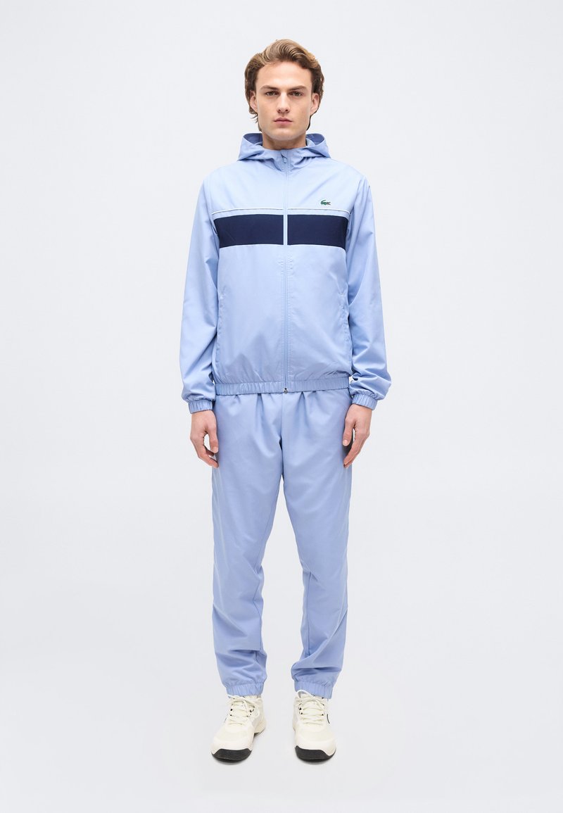 Νέος άνδρας που στέκεται φορώντας ένα ανοιχτό γαλάζιο φόρμα με ένα ναυτικό ρίγα στο στήθος και ανοιχτόχρωμα αθλητικά παπούτσια σε λευκό φόντο.