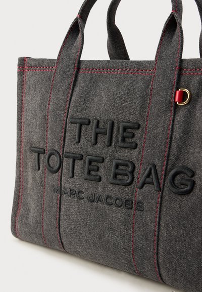Γκρι τσάντα tote από ύφασμα με κόκκινες ραφές, με ανάγλυφο κείμενο «THE TOTE BAG» και «MARC JACOBS», με χρυσές μεταλλικές λεπτομέρειες στο πλάι.