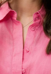Donna che indossa una camicia rosa con bottoni, una catena di collana d'argento visibile e capelli castano scuro sul lato destro.