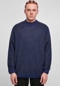 Tummansininen neulepaita, jossa on ribbikuvio, ylipitkä malli, korkea kaulus ja laskeutuvat hartiat. Varustettu pitkillä hihoilla ja rentoilla hihansuilla.