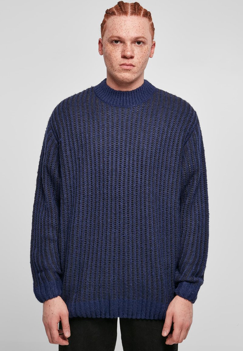 Tummansininen neulepaita, jossa on ribbikuvio, ylipitkä malli, korkea kaulus ja laskeutuvat hartiat. Varustettu pitkillä hihoilla ja rentoilla hihansuilla.