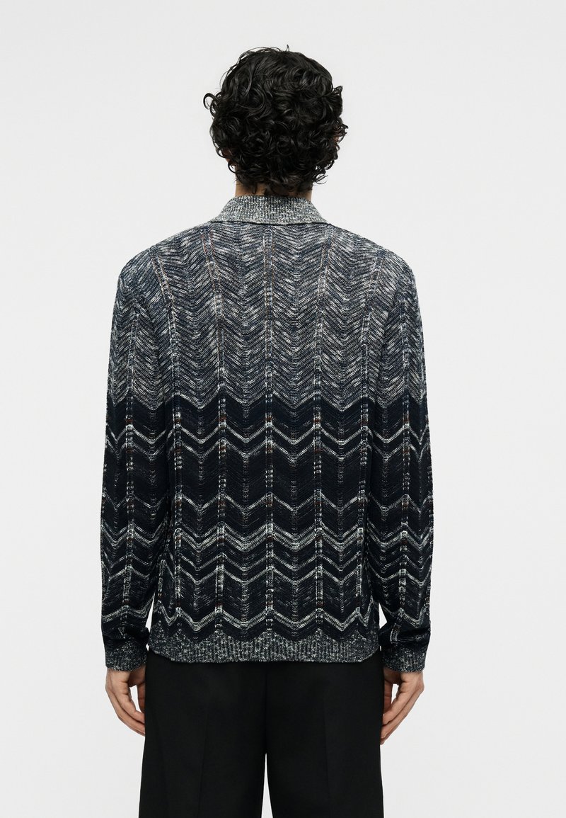 Man with curly hair wearing black and gray zigzag patterned knit sweater, shown from the back against a plain light background.