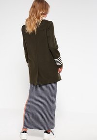 Abrigo de lana verde oliva con puños de manga a rayas blancas, combinado con una falda maxi a rayas azul marino y blanca con aberturas laterales y acentos rosas.