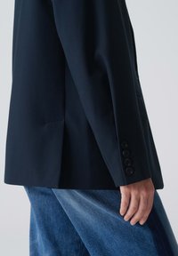 Blazer bleu marine avec des épaules structurées, une fente simple et des poignets à quatre boutons. Associé à un jean en denim bleu clair. Texture de tissu lisse.