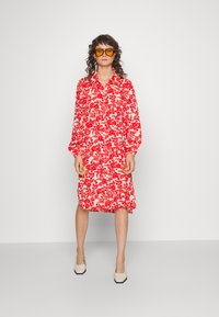 Vestido vermelho com flores, de mangas compridas e colarinho, feito de tecido leve; possui uma silhueta fluida e uma bainha até ao joelho. Usado com óculos de sol amarelos.