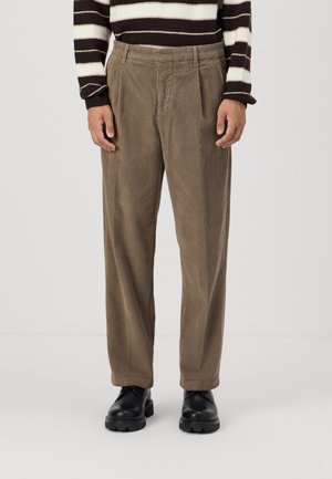 Pantaloni in corduroy marrone con una vestibilità ampia, caratterizzati da pieghe frontali e una superficie texturizzata. Abbinati a scarpe in pelle nere.
