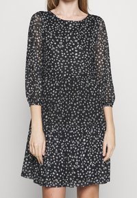 Kvinna iklädd en svart klänning med små vita blommönster, genomskinliga trekvartsärmar och en lager-på-lager kjol.