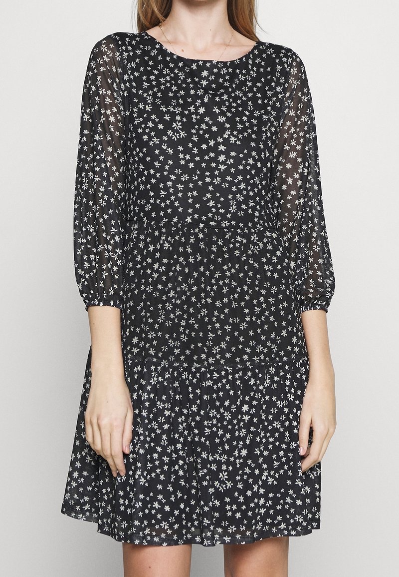 Kvinna iklädd en svart klänning med små vita blommönster, genomskinliga trekvartsärmar och en lager-på-lager kjol.