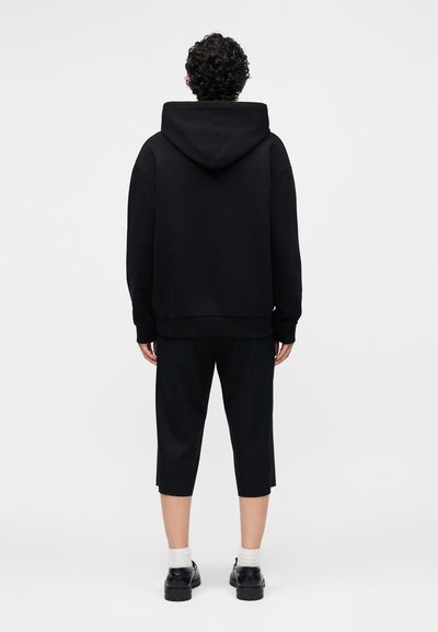 Άτομο με σγουρά μαλλιά που φοράει μαύρο φούτερ με κουκούλα, μαύρο κοντό παντελόνι, λευκές κάλτσες και μαύρα παπούτσια, στέκεται σε λευκό φόντο, από πίσω.