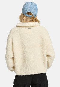 Cremfarbener Pullover mit Fleece-Textur und hohem Kragen. Verfügt über ein Etikett auf der Rückseite und hat eine lockere Passform, kombiniert mit einer hellblauen Jeans-Cap.