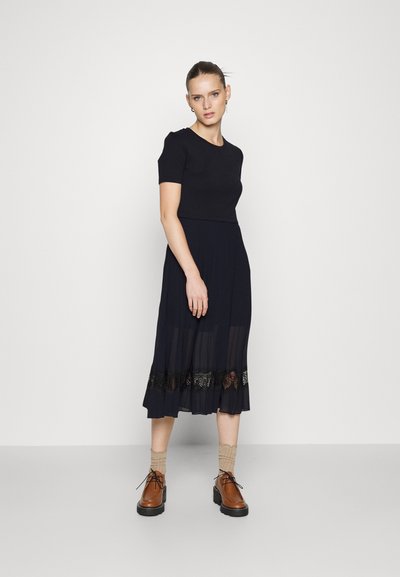 Vestido marino con un top ajustado y una falda plisada, que presenta un dobladillo transparente con detalles de encaje negro. Combinado con zapatos marrones y calcetines beige.