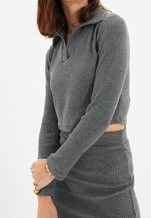 Frau trägt ein graues, geripptes Langarmoberteil und dazu passenden Rock, geschichtet mit goldenen Armbändern und Ringen an ihrer linken Hand.