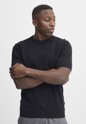 Black short-sleeve knit sweater with a crew neck, ribbed cuffs, and a textured fabric. The model's arms are crossed at the front.