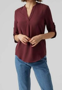Blouse bordeaux avec un col en V et des manches trois-quarts, fabriquée en tissu doux. Présente un ourlet arrondi et des détails de manches à rabat.