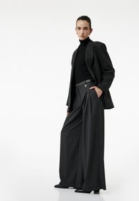 Sort blazer med struktureret stof lagdelt over en tætsiddende sort turtleneck. Bredbenede grå bukser med folder, stylet med et slankt sort bælte og hæle.