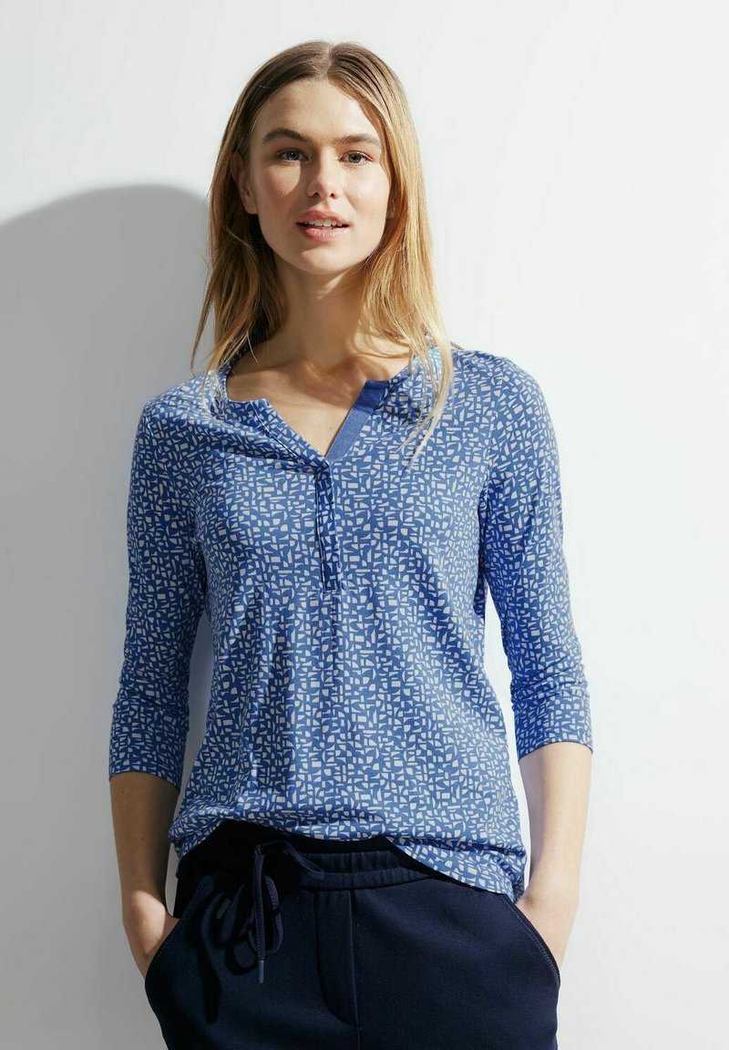 Niebieska koszula w wzory z rękawami o długości trzech ćwierci, z dekoltem w kształcie litery V i teksturowanym materiałem. Dodatki obejmują małą listwę z guzikami.