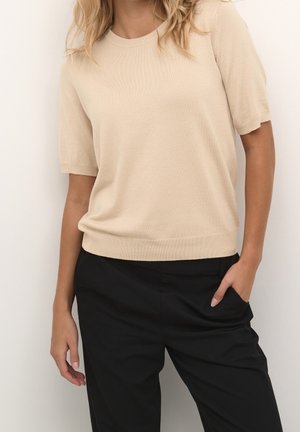 Vrouw met een beige gebreide top met korte mouwen en een zwarte broek, met één hand in de zak, staand tegen een lichte achtergrond.