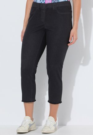 Femme portant un jean noir court à ourlet brut et des baskets blanches, se tenant contre un mur blanc.