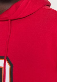 Rote Kapuzenpullover aus weichem Material, mit Kordelzug, einem großen Druckdesign in Weiß und Schwarz sowie kontrastierenden orangefarbenen Akzenten.