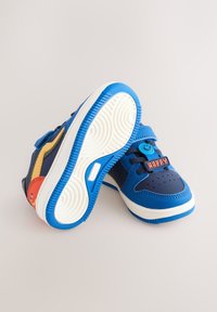 Baskets bleues avec un mélange de matériaux, dotées d'accents jaunes et rouges, d'un détail de visage souriant et d'une semelle blanche avec une bande de roulement texturée.