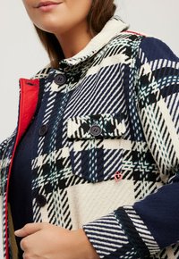 Giacca in misto lana con un motivo a quadri in blu navy, bianco e verde, dettagli rossi, due tasche sul petto e bottoni neri.