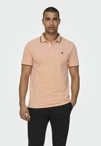 Camisa polo de color rosa claro con ribete negro en el cuello y las mangas, que presenta tres botones y un pequeño logo en el pecho.