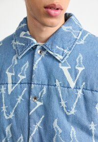 Primer plano de una persona que lleva una chaqueta de mezclilla azul con patrones de alambre de espino blancos y letras V, con botones a presión de metal y un cuello.