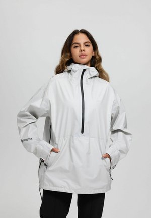 Junge Frau trägt eine weiße Gore-Tex wasserdichte Jacke mit Kapuze, Hände in den Seitentaschen, steht vor einem schlichten hellgrauen Hintergrund.