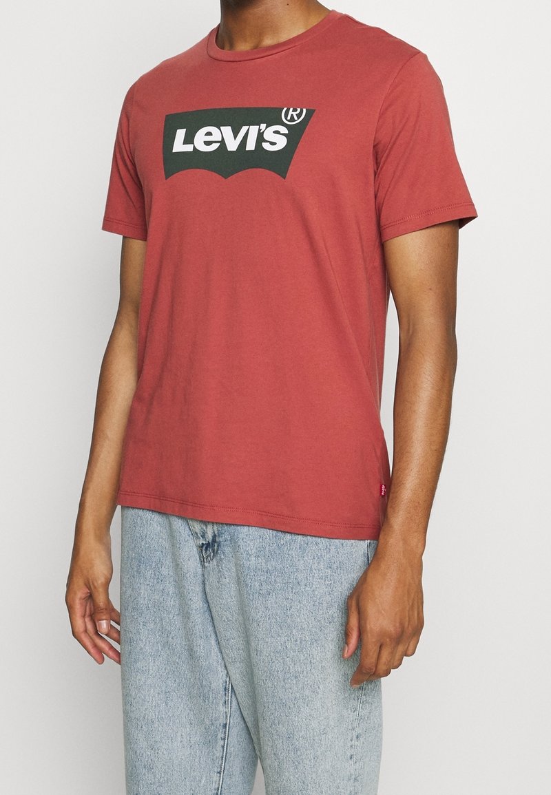 Personne portant un t-shirt Levi’s rouge rouille avec un logo noir et blanc, associé à un jean en denim bleu clair sur un fond uni.
