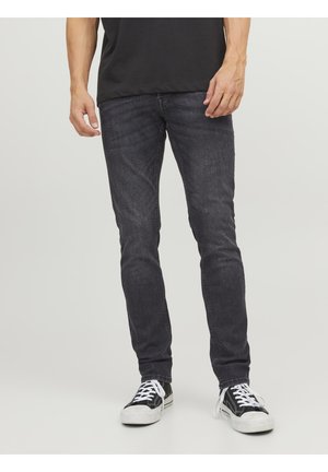 Mann trägt schwarze Slim-Fit-Jeans und schwarze Sneaker mit weißen Schnürsenkeln, steht vor einem schlichten hellen Hintergrund.