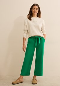 Sweatshirt crème avec le texte « il paradiso », associé à un pantalon vert ample avec taille à cordon, et des chaussures slip-on beiges.
