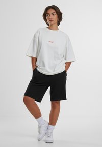 Tricou alb oversized cu text brodat roșu, combinat cu șorturi negre cu textură și adidași albi, având un material texturat și o croială lejeră.