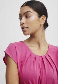 Fuchsia Bluse mit kurzen Ärmeln, die vertikale Falten um den Ausschnitt und eine glatte, leichte Textur aufweist.