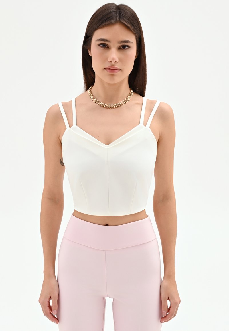 Jonge vrouw met lang donker haar, gekleed in een wit cropped topje met dubbele bandjes, lichtroze broek met hoge taille en een gouden ketting.