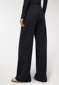 Pantaloni a gamba larga in un tessuto scuro, con vita alta, texture liscia e linee pulite, abbinati a scarpe slip-on.