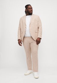 Helles beiges Leinenanzug mit einem taillierten Blazer und passender Hose, kombiniert mit einem weißen T-Shirt und weißen Sneakern. Glatte Textur, minimales Design.