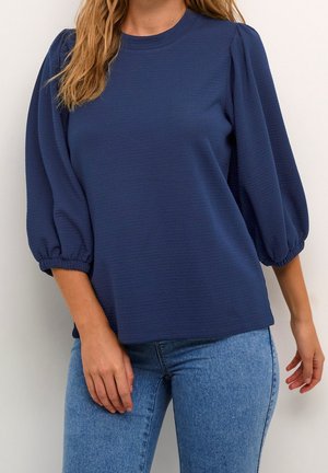 Vrouw met een marineblauwe blouse met pofmouwen en elastische manchetten en een lichtblauwe spijkerbroek, staand tegen een eenvoudige lichte achtergrond.