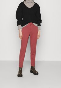 Zwarte trui met een losse pasvorm, gelaagd over een gestreepte coltrui. Rode slim-fit broek en bruine enkellaarsjes met een dikke zool.