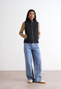 Črn prešiti jopič z visokim ovratnikom, v kombinaciji s svetlim dolgim rokavom in širokimi modrimi kavbojkami. Bele superge z rumenim podplatom.