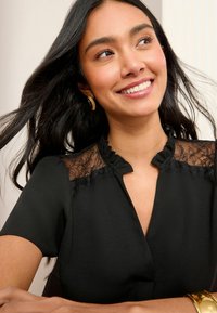 Blouse noire avec épaules en dentelle, col froncé et manches courtes. Tissu lisse avec un léger éclat, complété par un bracelet doré.