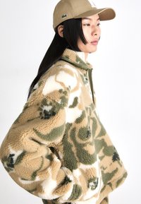 Veste en polaire beige, verte et blanche avec un motif camouflage. Dotée d'un col montant et d'une fermeture éclair. Portée avec une casquette beige.