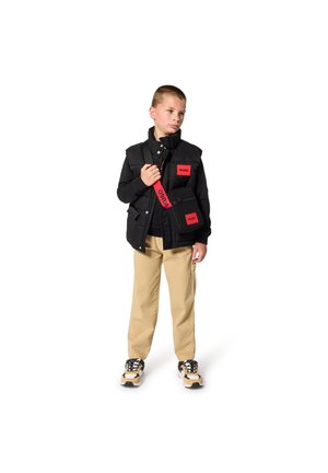 Gilet imbottito nero con etichette rosse, pantaloni beige e scarpe da ginnastica. Accessori comprendono una piccola borsa con tracolla rossa. Design semplice e funzionale.