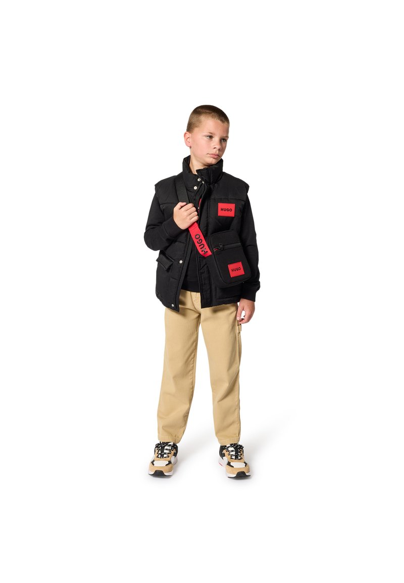 Μαύρο αμάνικο μπουφάν τύπου puffer με κόκκινες ετικέτες, μπεζ παντελόνι και αθλητικά παπούτσια. Αξεσουάρ περιλαμβάνουν μια μικρή τσάντα με κόκκινο λουρί. Απλός, λειτουργικός σχεδιασμός.